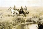 Three Piegan men on horseback in open grassland near pond.
